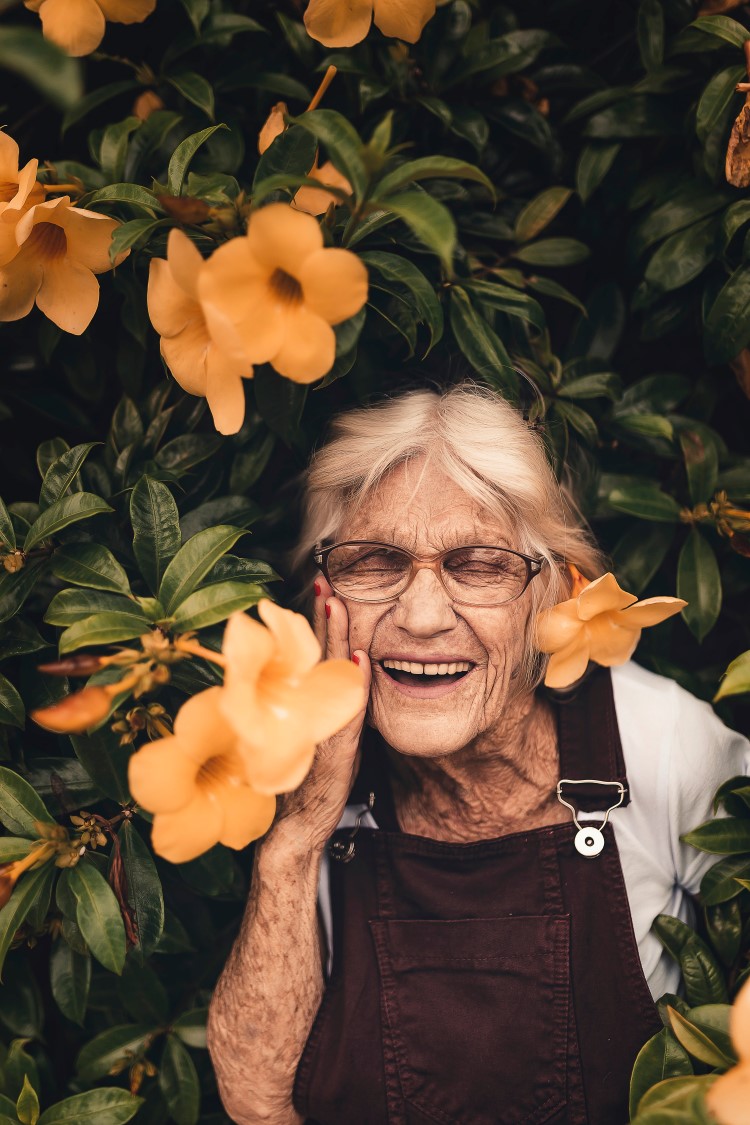 Eldre dame som står inntil et tre med blomster